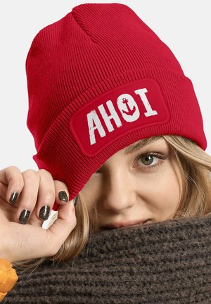 Red knit beanie with a rectangular patch featuring the word "AHOI" and an anchor design. Ribbed texture with a snug fit.