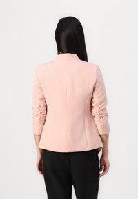 Pink tailored blazer with long sleeves, featuring gathered detailing at cuffs and a high collar. Soft fabric with a smooth texture.