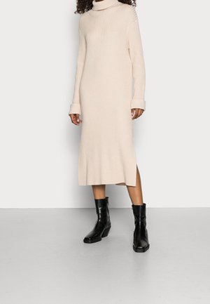 Žena v béžových žebrovaných midi šatech s rolákem a dlouhými rukávy, v černých kotníkových botách, stojící na šedé podlaze.