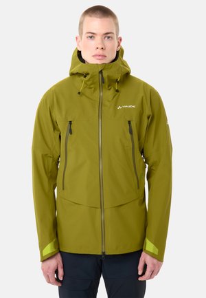 Jonge persoon met kort haar die een olijfgroene Vaude waterdichte jas draagt met een voorrits en capuchon, staand tegen een effen lichte achtergrond.