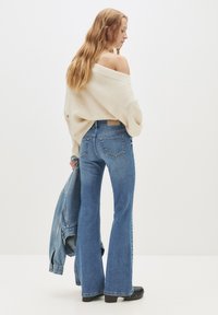 Nainen, jolla on pitkät hiukset, pukeutuneena off-shoulder-kermaiseen neuleeseen, sinisiin leveälahkeisiin farkkuihin ja mustiin puukenkään, joka pitää denim-jakkuja valkoista taustaa vasten.