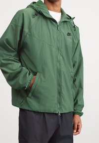 Green hooded jacket with a zipper, made from lightweight material. Features side pockets and black accents on the zippers and cuffs.