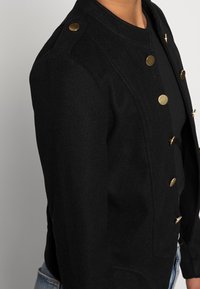Giacca nera in misto lana con scollatura rotonda, bottoni dorati e spalle strutturate. Presenta tasche frontali e una vestibilità sartoriale.