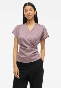 Blusa a wrap in morbido satin, colore malva, con maniche corte e dettagli di drappeggio leggeri, abbinata a pantaloni neri.