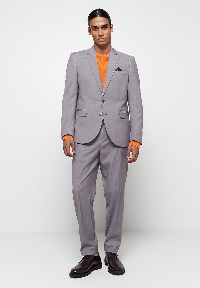 Lichtgrijs pak met een slim fit, met een enkelvoudige sluiting, notch lapels en twee voorzakken. Gekozen over een oranje trui.