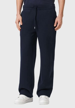 Mand iført løse marineblå sweatpants med snøre og hvide sneakers, stående med hænderne afslappet ned langs siderne.