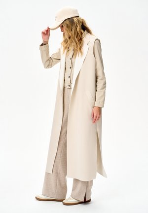 Kobieta w beżowej długiej kamizelce, dopasowanych spodniach, zapiętej koszuli, trampkach i czapce, pozująca z pochyloną głową i ręką poprawiającą czapkę.