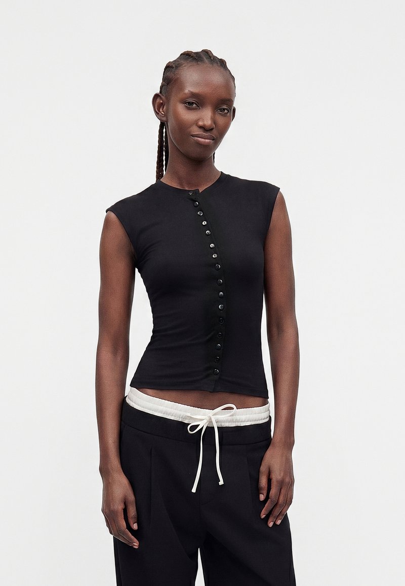 Femme aux cheveux tressés, portant un haut noir sans manches boutonné et un pantalon noir avec une taille à cordon blanc, sur un fond uni.