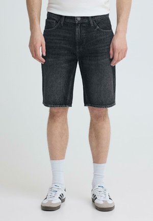 Mann iført svarte denimbukser, hvite crew-sokker og hvite sneakers, stående mot en ensfarget bakgrunn med hendene avslappet langs siden.