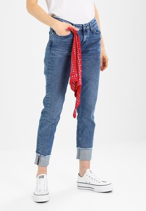 Persona che indossa jeans blu risvoltati e sneaker bianche, che tiene in vita un bandana rosso con motivi di stelle e punti bianchi.