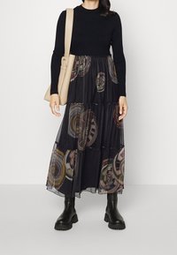 Maglia nera a maniche lunghe con costine abbinata a una gonna maxi nera trasparente a strati con motivi a spirale colorati. Indossata con stivali neri alti e una borsa leggera.