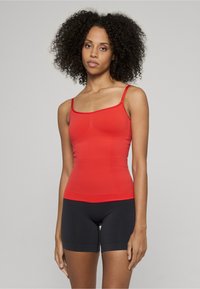 Rød tettsittende tanktop med tynne stropper, sømløst design og elastisk stoff, kombinert med svarte shorts til midten av låret med glatt tekstur.