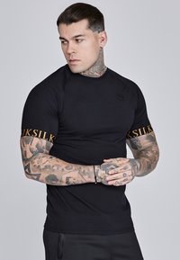 Schwarzes kurzärmliges Shirt aus dehnbarem Material mit rundem Ausschnitt und goldener "SikSilk"-Druck auf den Ärmeln.