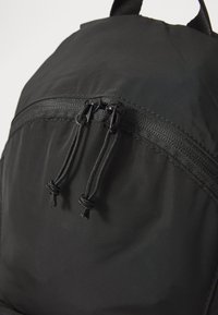 Fekete hátizsák sima nylon külsővel, cipzáras zsebbel és zsinóros záró mechanizmussal. Texturált elemek a cipzár közelében.