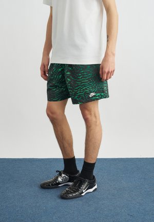 Muž v bielom tričku, čiernych a zelených kraťasoch Nike s vzorom, čiernych ponožkách a čiernych teniskách Nike na modrom koberci.