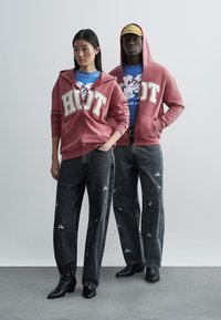 Duas pessoas estão juntas vestindo roupa casual. Uma veste um hoodie com fecho em tom bordô com a palavra "HOT" em branco e jeans pretos com padrão floral. A outra usa uma t-shirt azul com gráfico de um personagem, combinada com calças pretas e botas.