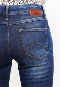 Jeans de mezclilla azul oscuro con un acabado texturizado, que presentan una etiqueta de cuero marrón con la inscripción 'Mustang' y un distintivo pespunte en los bolsillos traseros.