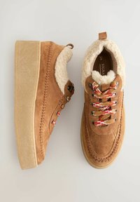 Tan suede sneakers with a faux fur lining, thick beige rubber soles, multicolored braided laces, and decorative stitching along the edges.