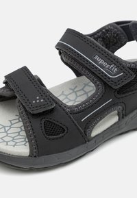 Sorte sandaler til småbarn i svart, med justerbare stropper, mesh-detaljer, gummisåle og en lys grå teksturert fotbed med geometrisk mønster.