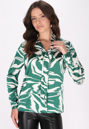 Grøn- og hvidmønstret bluse med knytkrave, lange ærmer og knapmanchetter. Stoffet har en glat tekstur. Båret sammen med sorte bukser.