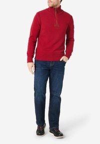 Roter Strickpullover mit hohem Kragen, Reißverschluss und gerippten Bündchen sowie Saum, kombiniert mit blauen Jeans und schwarzen Schnürschuhen.