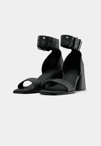 Schwarze High Heel Sandalen mit einem Knöchelriemen und silbernen Beschlägen, aus glattem Leder, mit offenem Zehenbereich und Blockabsatz-Design.