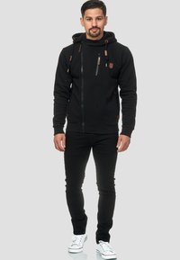 Sudadera negra con cremallera y cuello alto, con acentos marrones y cordones. Combinada con pantalones negros ajustados y zapatillas blancas.