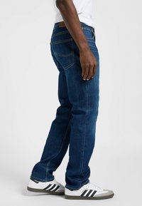 Jeans de mezclilla azul oscuro, corte recto, diseño de cinco bolsillos, con detalles de costura amarilla y un parche trasero de cuero marrón. Llevados con zapatillas blancas.