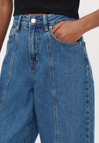 Jeans in denim blu a vita alta, caratterizzati da una vestibilità rilassata, cuciture dorate, tasche frontali e una chiusura a bottoni in vita.