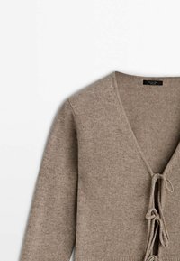 Beige neulehuppari, jossa on V-kaula-aukko, pitkät hihat ja edessä solmittava kiinnitys. Pehmeä rakenne, jossa on vähän yksityiskohtia.