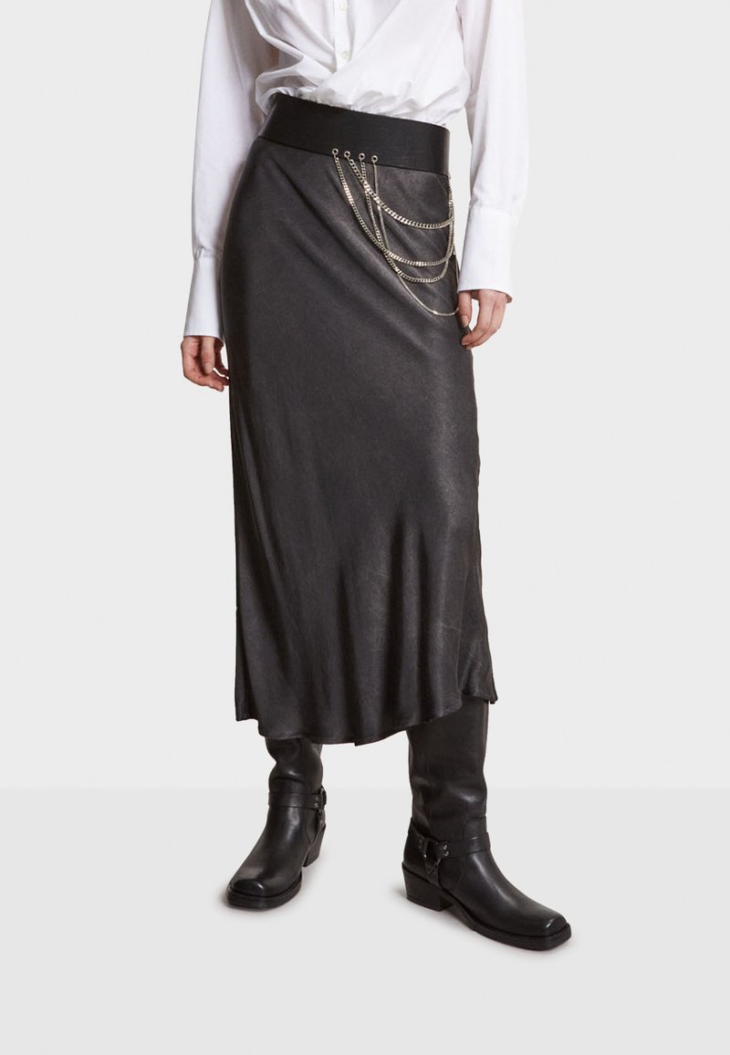 Jupe noire longue jusqu'à la cheville en matériau lisse, avec une large ceinture et un embellissement en chaînes argentées superposées au niveau des hanches.