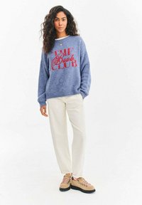 Niebieski puszysty sweter z czerwonym napisem "THE Book CLUB", zestawiony z jasnokremowymi spodniami i beżowymi botkami z różowymi sznurowadłami i czarnymi podeszwami.