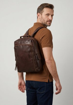 Man draagt een bruin poloshirt en spijkerbroek, draagt een grote donkerbruine leren rugzak en kijkt over zijn schouder tegen een effen achtergrond.
