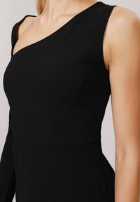 Musta mekko epäsymmetrisellä kaula-aukolla, istuvalla bodysuitilla ja yhdellä pitkällä hihalla. Sujuva pinta ja yksinkertainen muotoilu ilman näkyviä kuvioita.