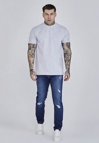 Svetlo siva kratka majica z okroglim ovratnikom, kombinirana s temno modrimi raztrganimi kavbojkami in belimi supergami. Tetovaže so vidne na rokah.