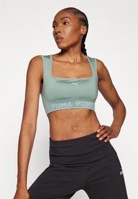 Šviesiai žalias sportinis liemenukas su kvadratine iškirpte, tekstūruota juosta su ryškiu užrašu "PUMA", derinamas prie juodų dideliu liemeniu kelnaičių.