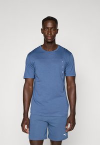 Camiseta atlética azul de tejido suave, con mangas cortas y un acento horizontal, y un logotipo impreso en vertical en el costado.