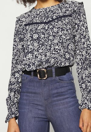 Blusa floreale navy con stampa botanica bianca, maniche arruffate e una cintura nera con fibbia rettangolare dorata abbinata a jeans in denim.