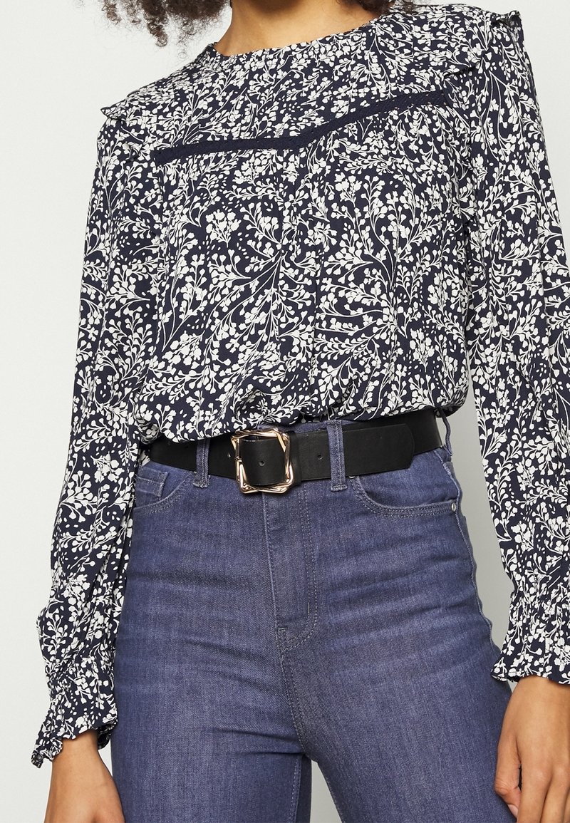 Blusa floreale navy con stampa botanica bianca, maniche arruffate e una cintura nera con fibbia rettangolare dorata abbinata a jeans in denim.