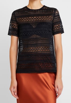 Femme portant un haut en dentelle noire à manches courtes avec des motifs géométriques sur la peau, associé à une jupe en satin couleur rouille.