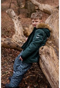 Green insulated jacket with a fur-lined hood, paired with blue patterned pants, sitting on a log in a forest setting.