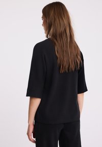 Kobieta z długimi brązowymi włosami, ubrana w czarny sweter o krótkim rękawie i czarne spodnie, stojąca bokiem na jednolitym tle.