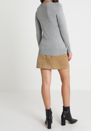 Femme debout, portant un haut gris à manches longues, une jupe en velours côtelé beige et des bottines à talons noires, de dos contre un fond uni.