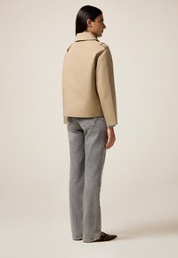 Beiger Trenchcoat mit strukturiertem Design, leicht verkürzter Länge und weiten grauen Denim-Hosen. Glatte Textur, minimaler Hardware, klare Linien.