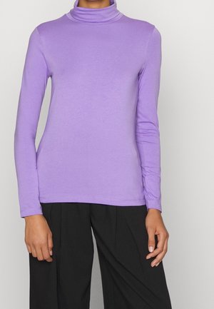 Personne portant un haut à col roulé ajusté lavande à manches longues et un pantalon noir ample sur fond uni.