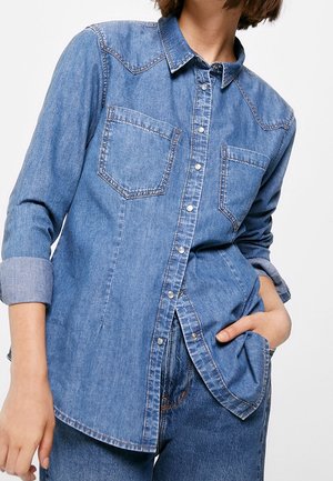 Camicia di jeans in azzurro chiaro, con due tasche sul petto, chiusura a bottoni e maniche arrotolate. Texture morbida con dettagli di cucitura a contrasto.