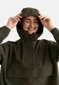 Femme ajustant la capuche d'une veste imperméable vert foncé, souriant les yeux fermés devant un fond uni.