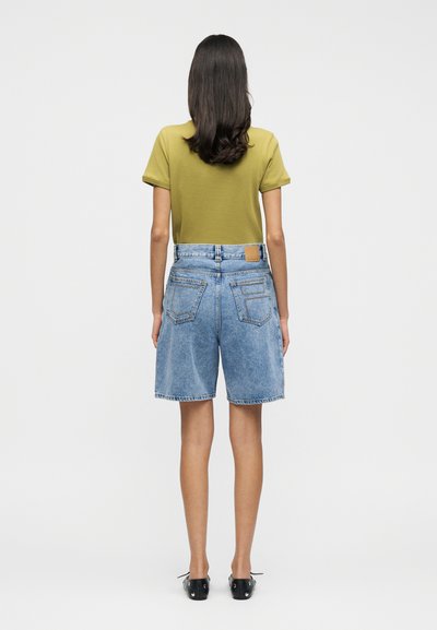 Ελιά πράσινο κοντομάνικο τοπ σε συνδυασμό με ανοιχτό μπλε τζιν σορτς με τετράγωνο πατς και δύο πίσω τσέπες, φορεμένα με μαύρα παπούτσια.