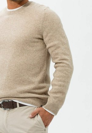 Homme portant un pull en maille beige, un sous-vêtement blanc, un pantalon beige clair et une ceinture marron avec la main dans la poche, sur un fond uni.