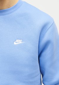 Ανοιχτό μπλε φούτερ με μικρό λευκό λογότυπο Nike κεντημένο στο αριστερό στήθος, φορεμένο από ένα άτομο μπροστά σε απλό φόντο.